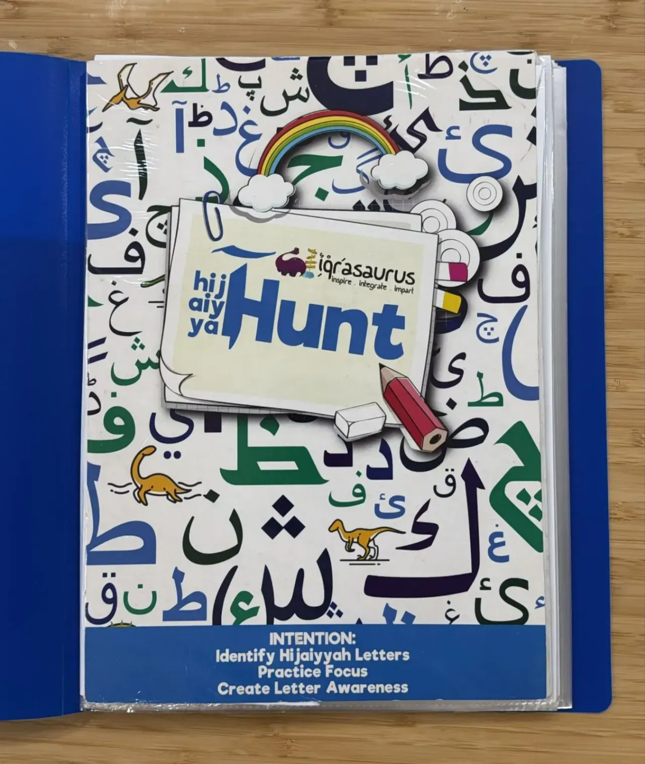 The printed Hijaiyya Hunt book in a blue folder, its cover full of Arabic and Jawi letters with small dinosaurs