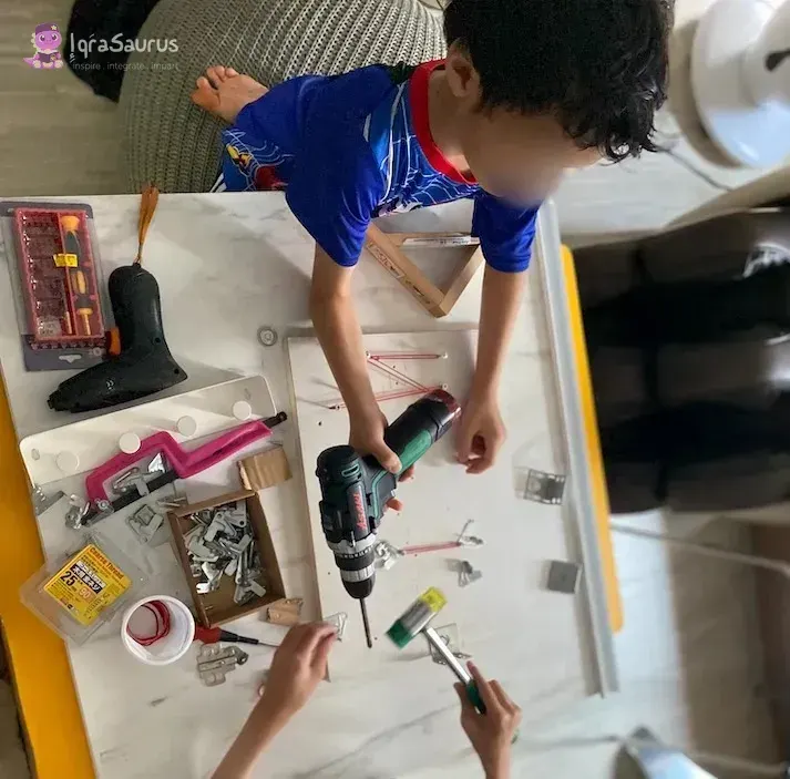 Seen from above, a preschooler uses a cordless drill on a board while an older helper holds a screwdriver