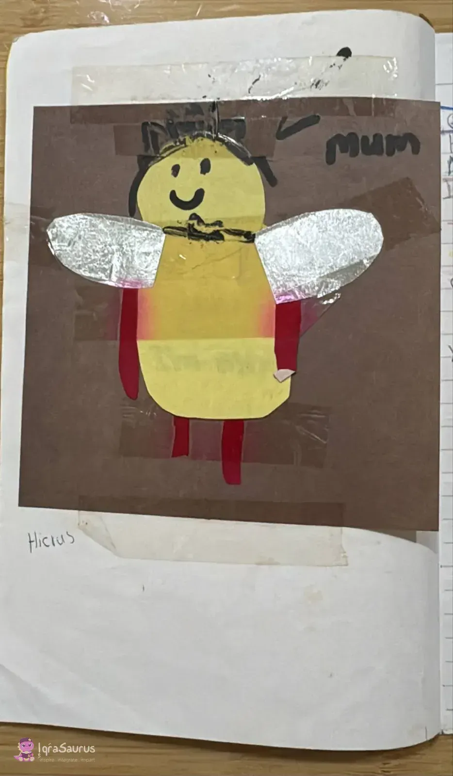A collage bee with a smiling face, foil wings and red legs, taped onto brown paper and labelled mum.