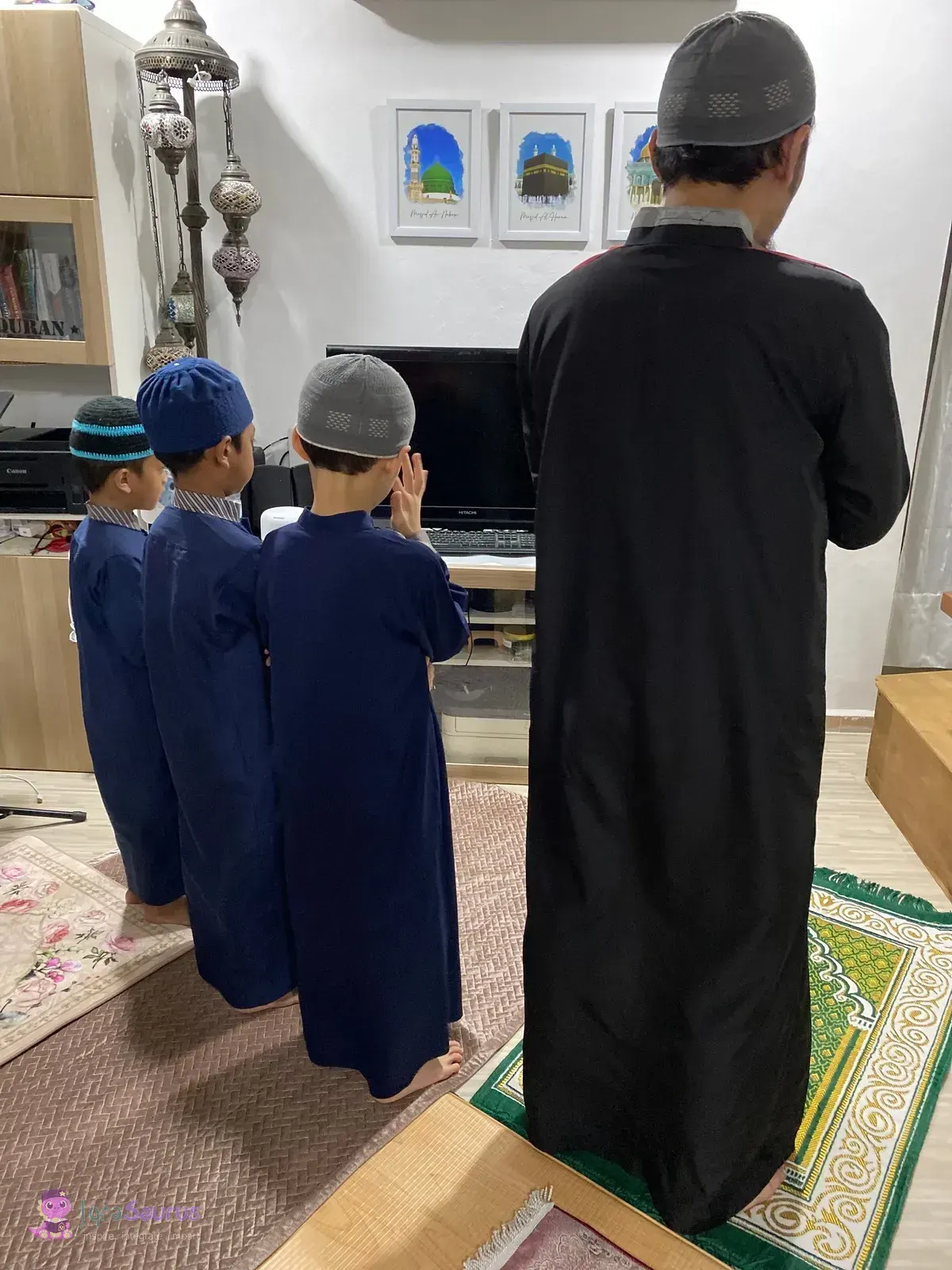 Three boys in navy jubah and kopiah pray in a row behind an adult leading the prayer at home